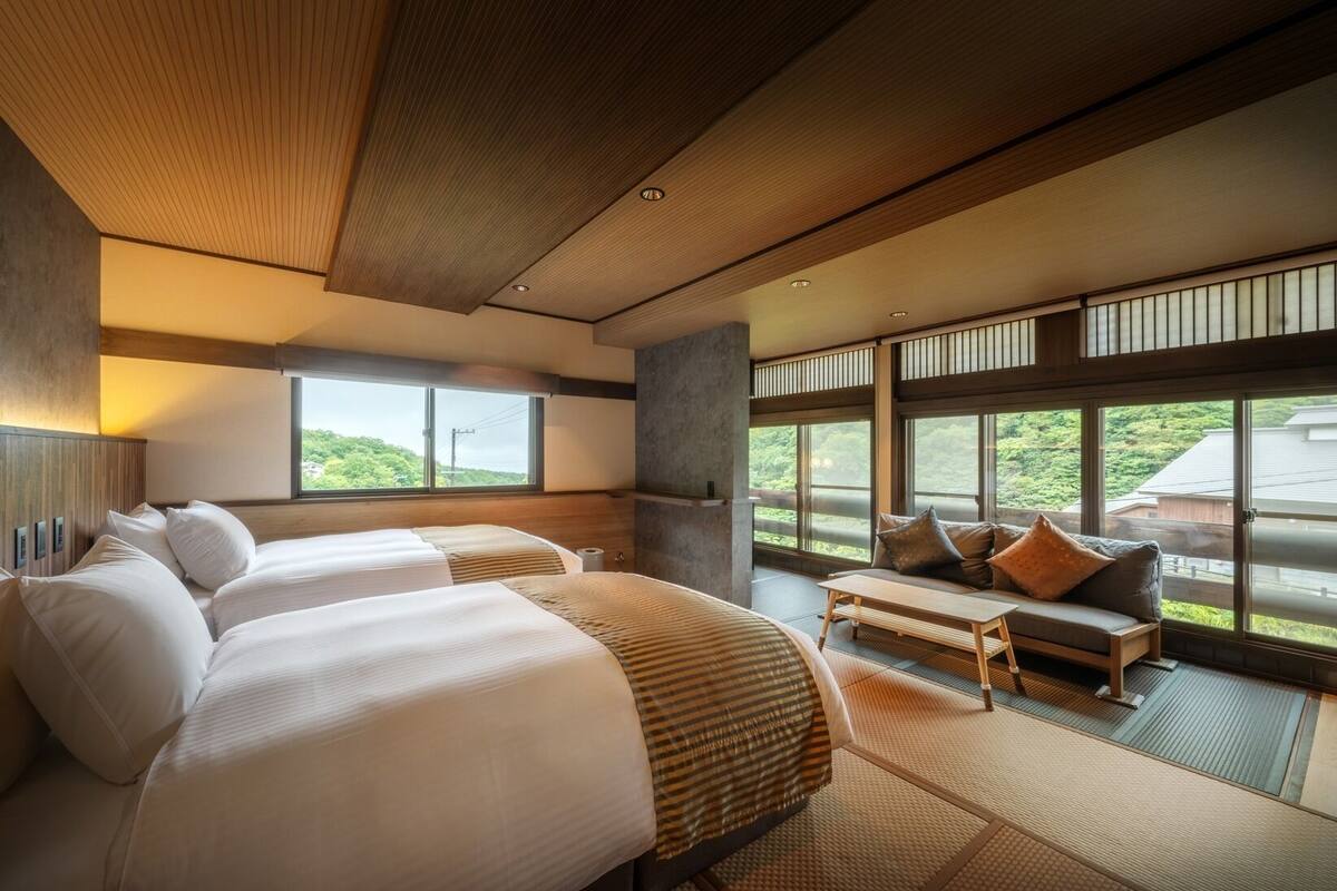 A hotel room with two beds, a sofa, a small table, and large windows offering a view of greenery.