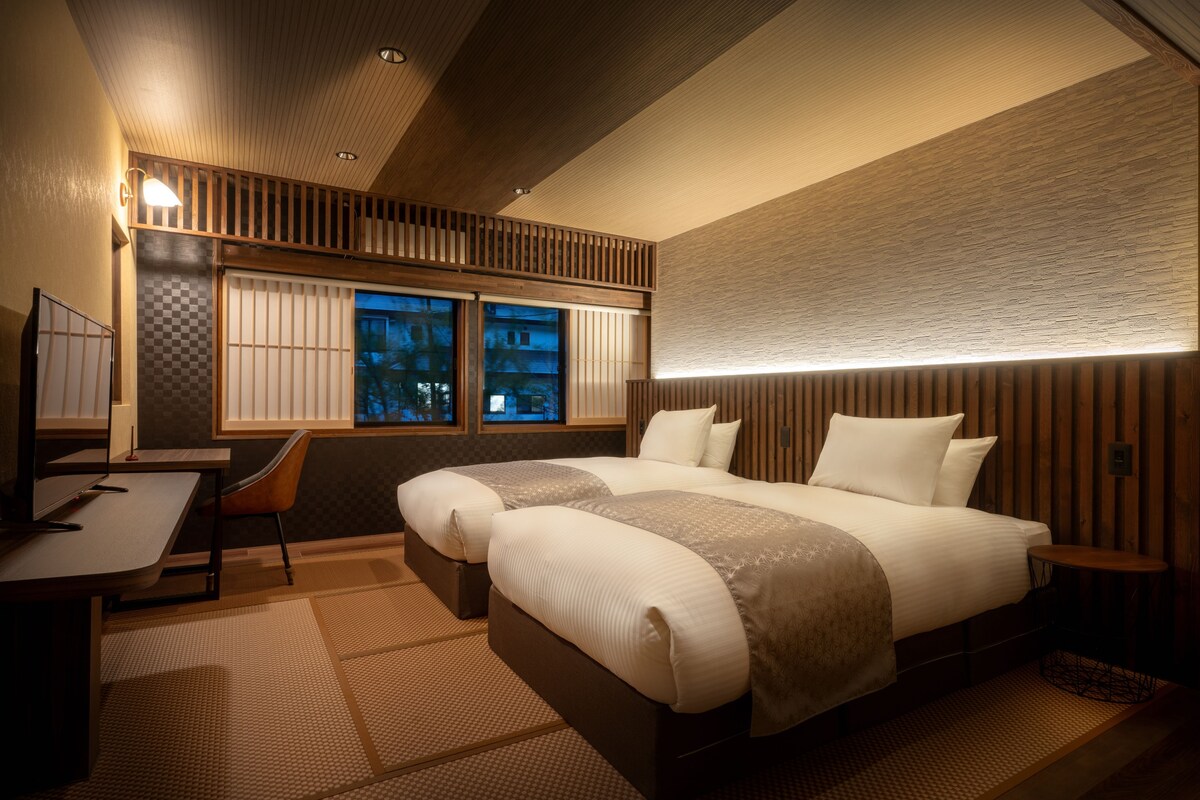 A hotel room with a large bed, a desk, and a chair. There are windows with a view of the outside.