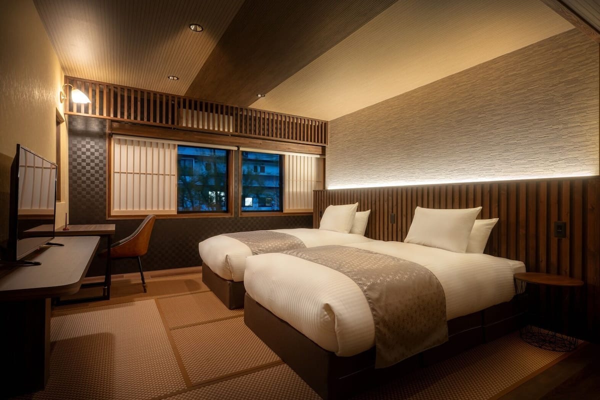 A modern hotel room with a large bed, a desk, and a view of the cityscape through the windows.