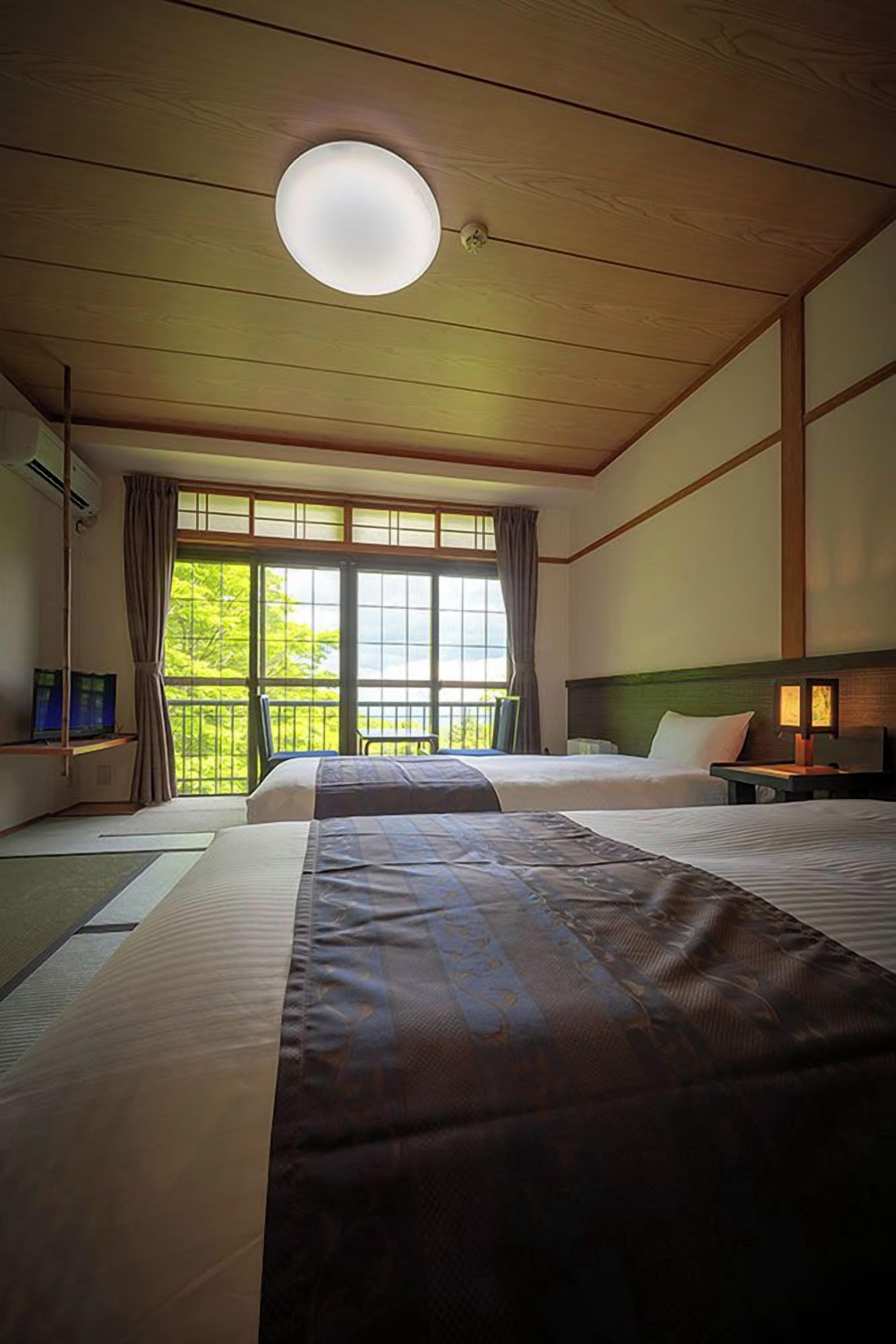 Quarto de hotel com duas camas, uma janela grande, uma luminária no teto e uma TV sobre um suporte.