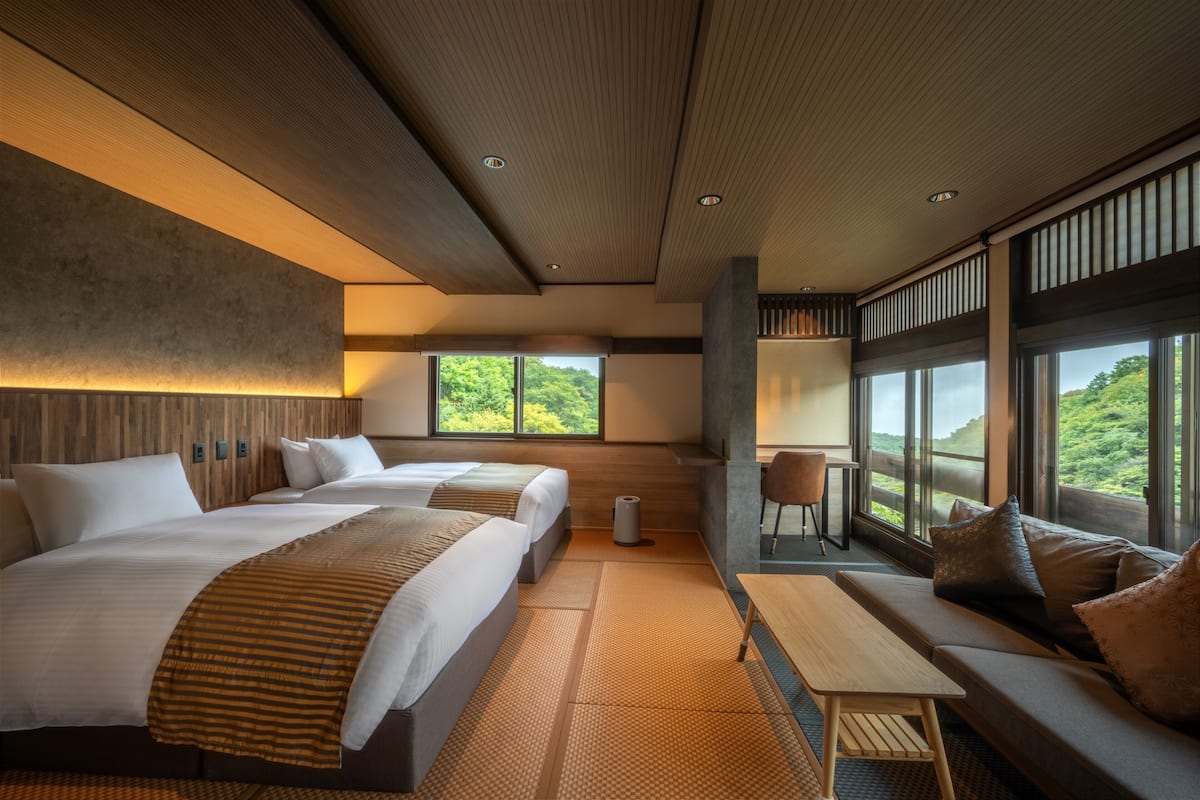 A modern hotel room with two beds, a sofa, a table, and large windows with a view of greenery.