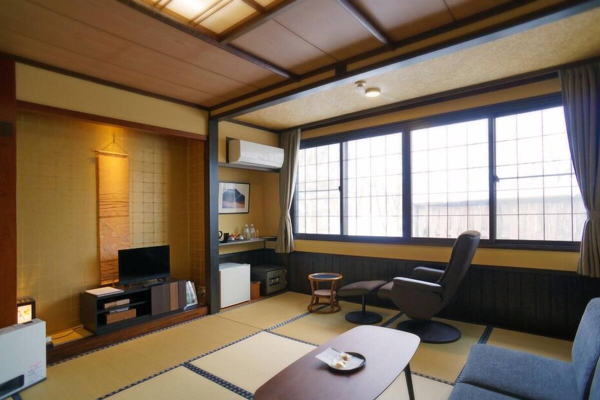 A traditional Japanese-style room with tatami flooring, a television, a sofa, a small table, and a window with curtains.