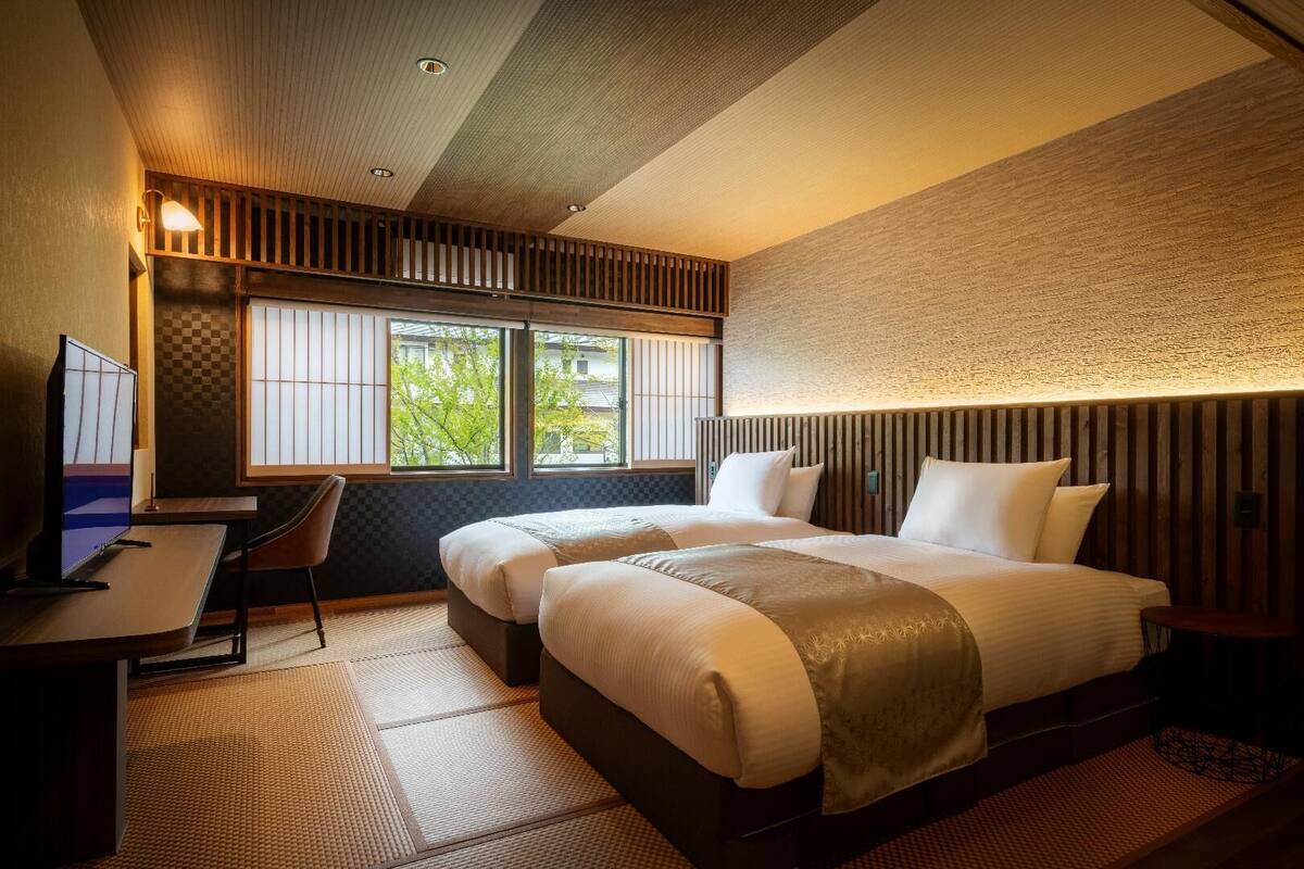 A hotel room with two beds, a desk, a chair, and a large window with a view of greenery.