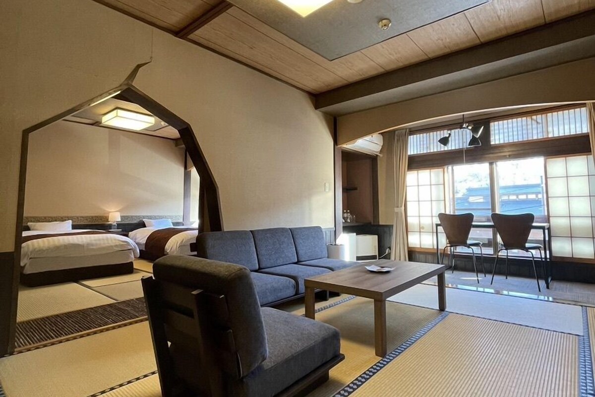 A hotel room with a sofa, a small table, a bed, and a view of the outside through large windows.