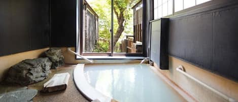 A traditional Japanese onsen with a stone bath, wooden benches, and a view of a tree through a sliding door.