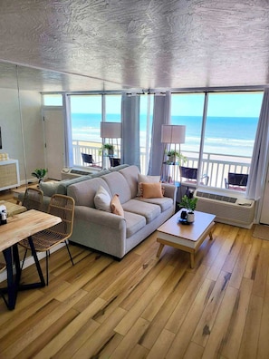 A modern living room with a large sofa, dining table, and chairs. It features large windows with a view of the ocean.
