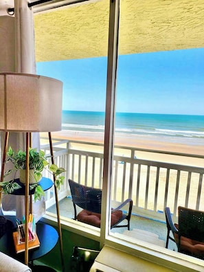 A room with a view of the beach, a lamp, a chair, and a table with a plant and stationery.