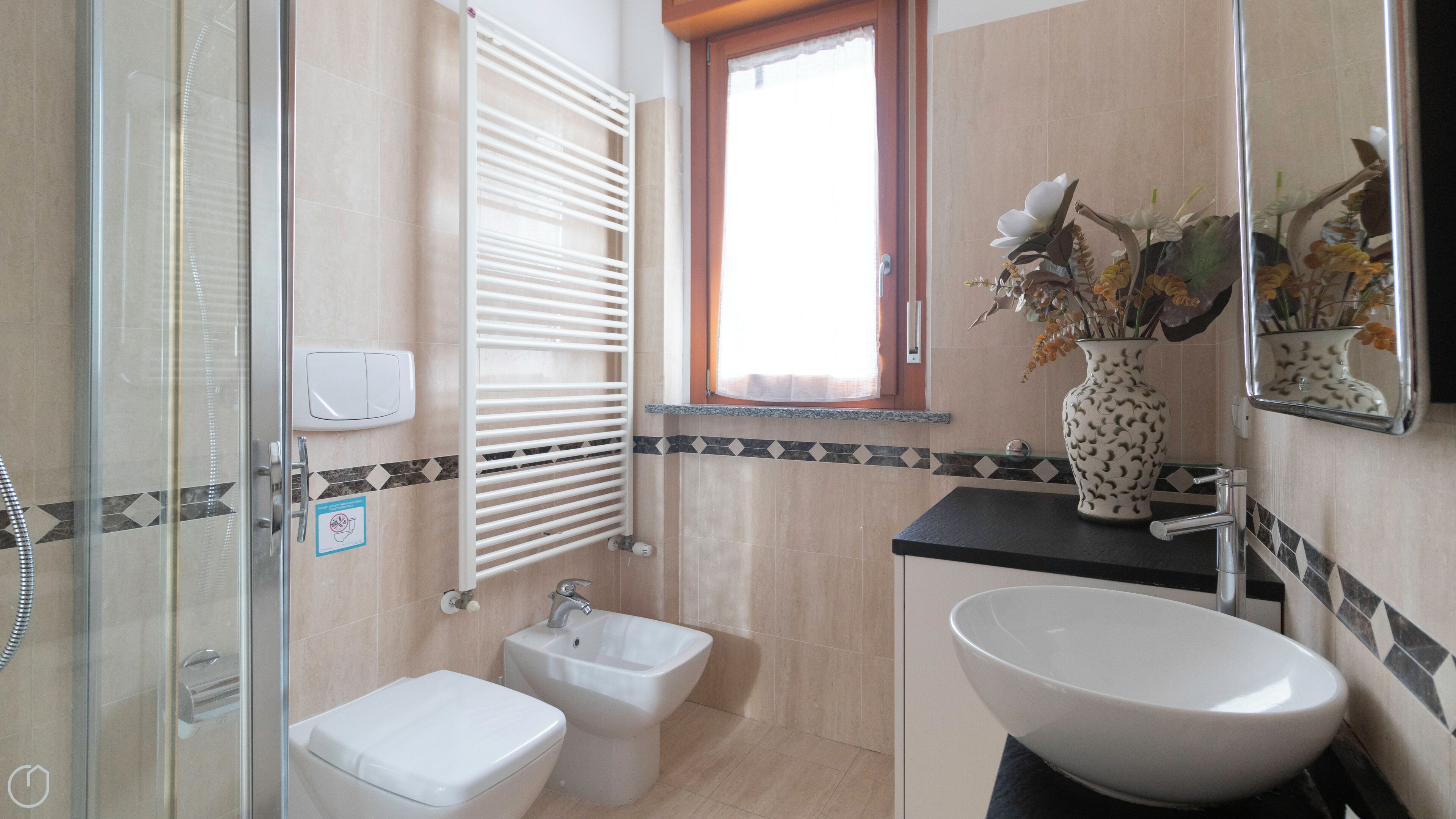 A bathroom with a glass shower enclosure, a bidet, a sink with a large basin, and a window with blinds.