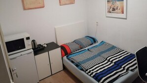 A small, neatly organized bedroom with a single bed, a desk with a computer, a microwave, and a small cabinet. There are two framed pictures on the wall. - All you need a comfy place (Amsterdam)