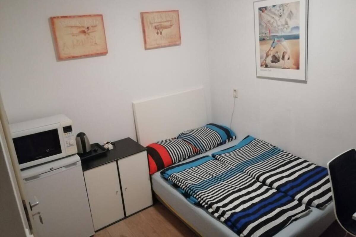 A small, neatly organized bedroom with a single bed, a desk with a computer, a microwave, and a small cabinet. There are two framed pictures on the wall.