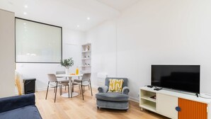 Un salon moderne avec un téléviseur à écran plat, un fauteuil gris, une table à manger avec des chaises et un meuble blanc avec des étagères.