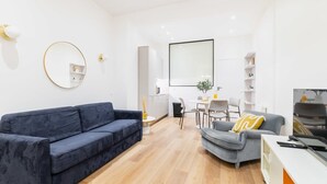 Un salon moderne avec un canapé bleu, un fauteuil gris et une table basse blanche. On y trouve également un coin repas avec une table et des chaises blanches, ainsi qu’une cuisine avec des placards blancs et une poubelle noire.