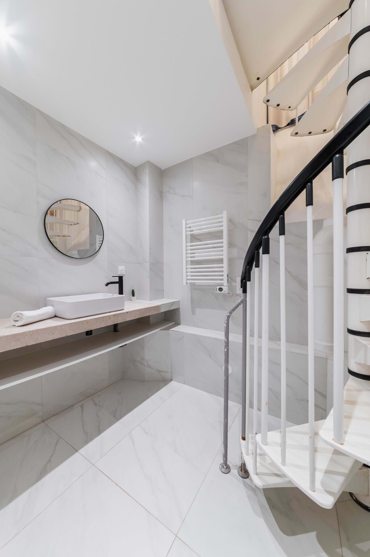 A modern bathroom with a white marble floor, a round mirror, a white sink, and a black and white staircase.