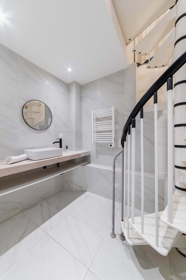 Un baño moderno con piso de mármol blanco, un espejo redondo, un lavamanos blanco y una escalera negra y blanca.