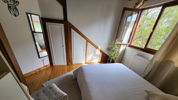 A bedroom with a large bed, wooden flooring, a small seating area, and a window with a view of greenery. - Charming chalet-style accommodation, 12 minutes from the town center (Schiltigheim)
