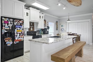 A kitchen with white cabinetry, a black refrigerator with various magnets, a wooden bench, and a marble countertop.