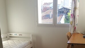 Ein Zimmer mit Holzboden, einem weißen Bett, einem Holztisch und einem Fenster mit Blick auf Gebäude.