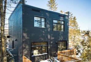 Un bâtiment moderne à deux étages, doté de grandes fenêtres, d’une terrasse en bois et surplombant un paysage enneigé.