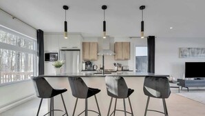 Una cocina moderna con una isla central, taburetes de bar, iluminación colgante y un amplio ventanal.