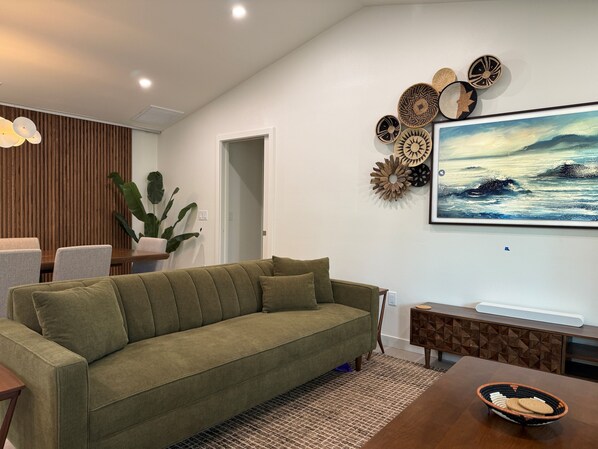 A modern living room with a green sofa, a dining table with a basket, and a wall with decorative plates and a painting.