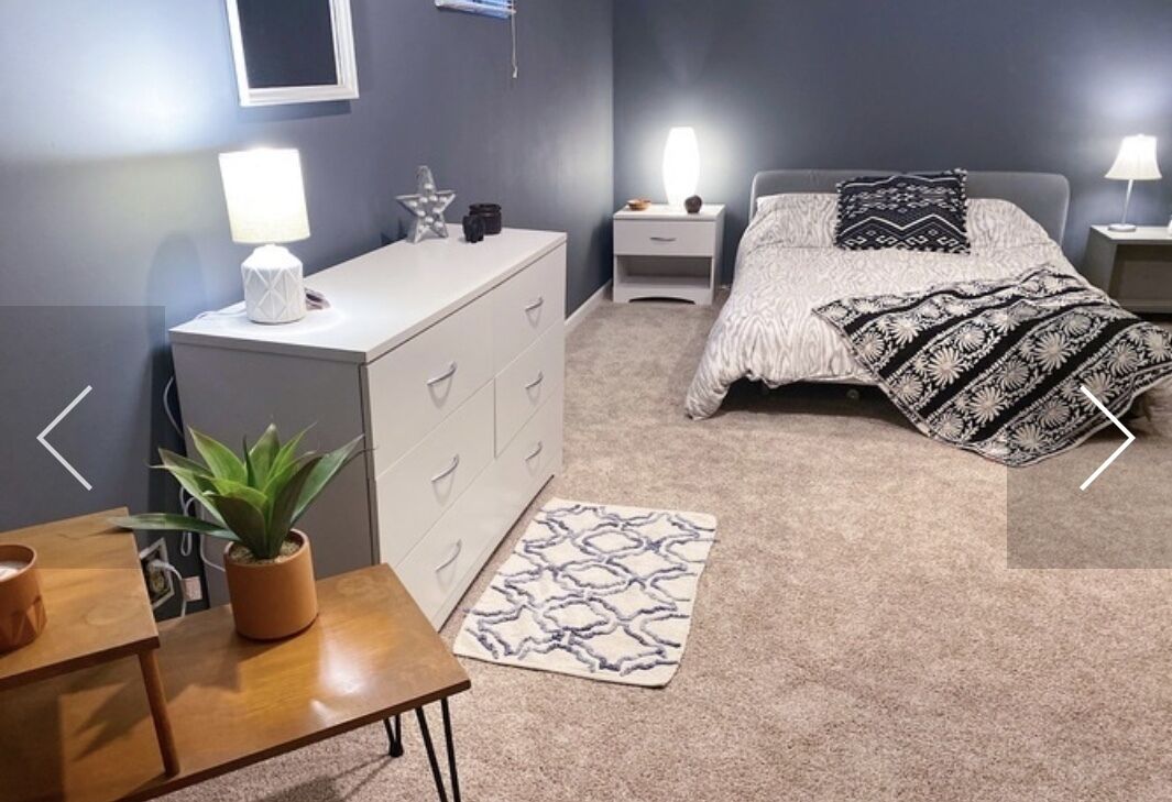 Ein Schlafzimmer mit Bett, Nachttischen, einem Schminktisch, einer Lampe, einer Pflanze und einem gemusterten Teppich.