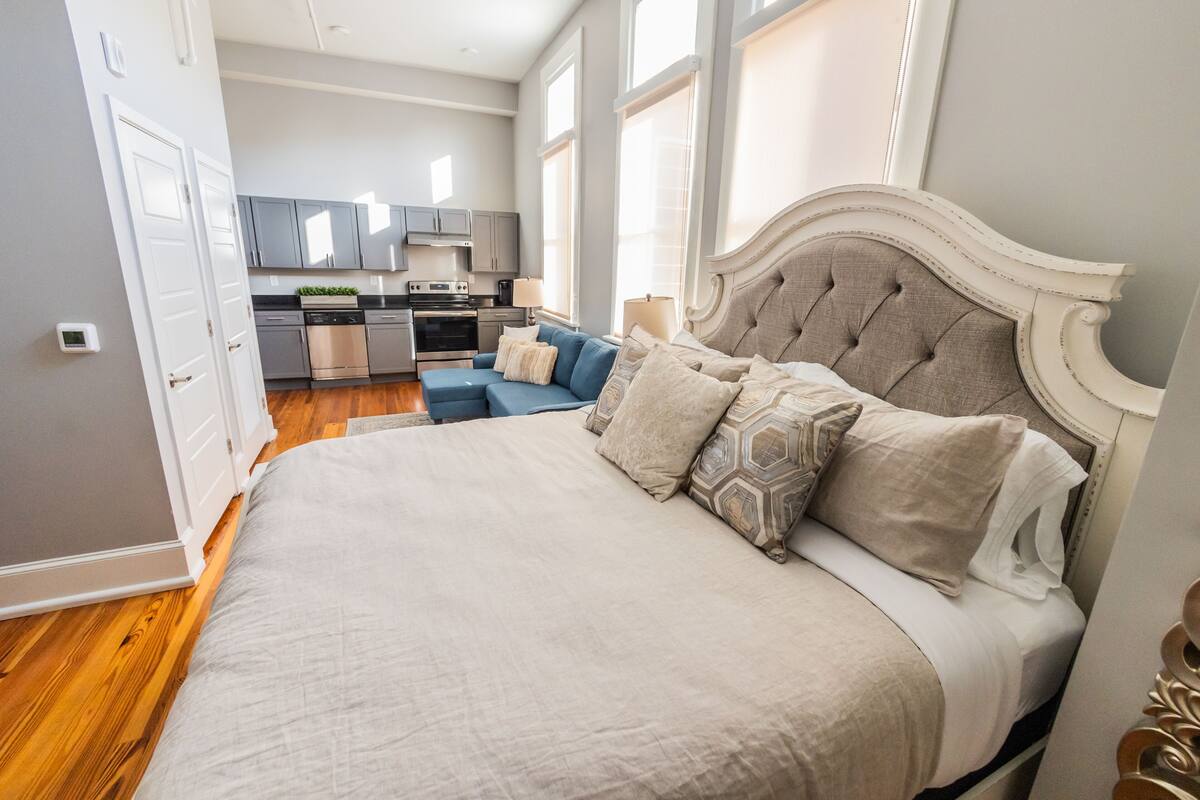 Quarto com uma cama grande, um sofá azul e uma cozinha ao fundo.
