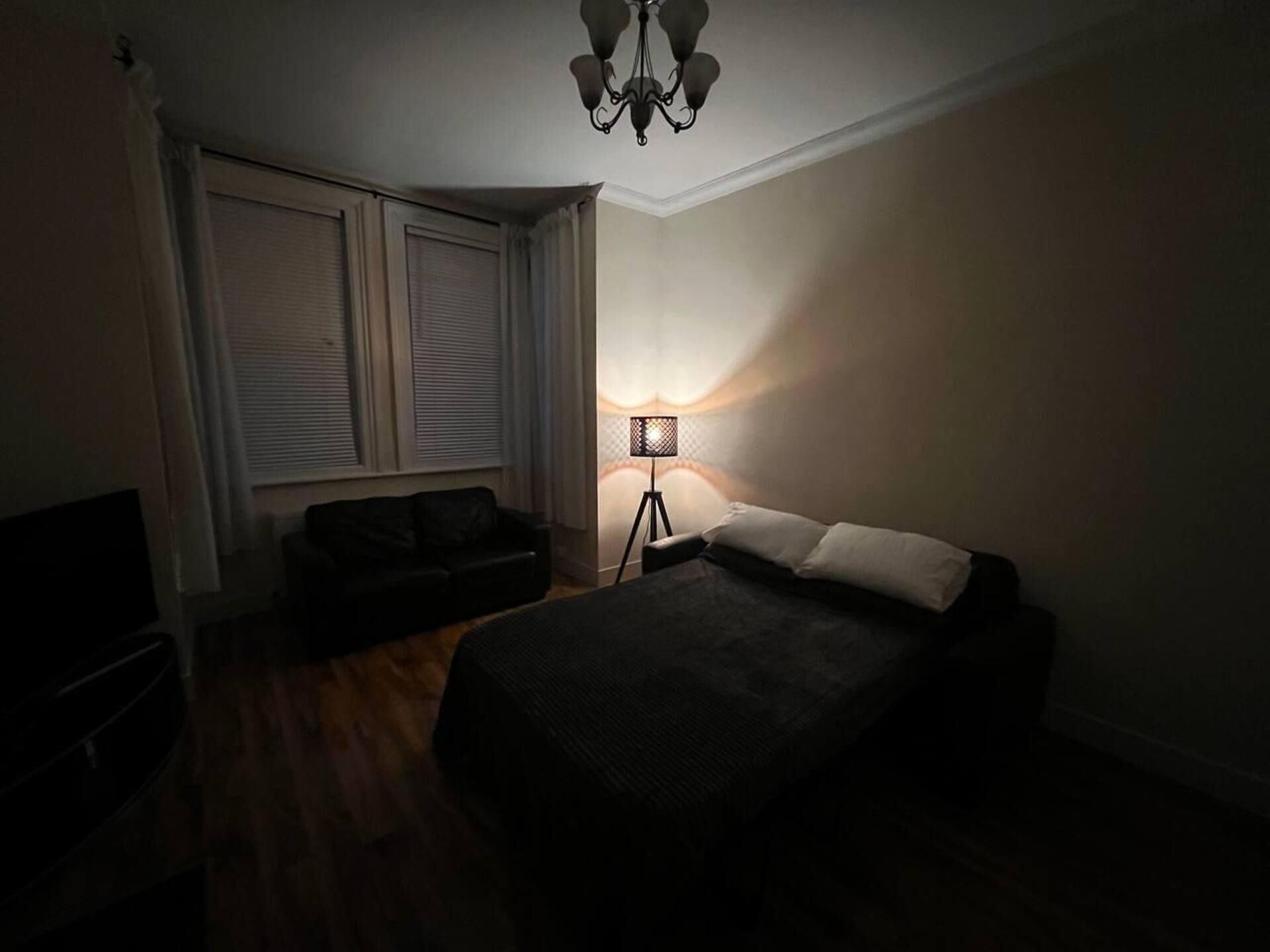 A bedroom with a bed, a standing lamp, a TV, and a window with blinds.
