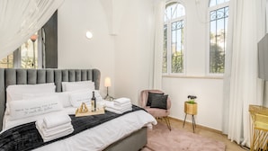 A neatly made bed with pillows, a bedside lamp, a small table with a bottle and glasses, a pink chair, and a window with a view. - Nina Luxury Apartment (Jerusalem)