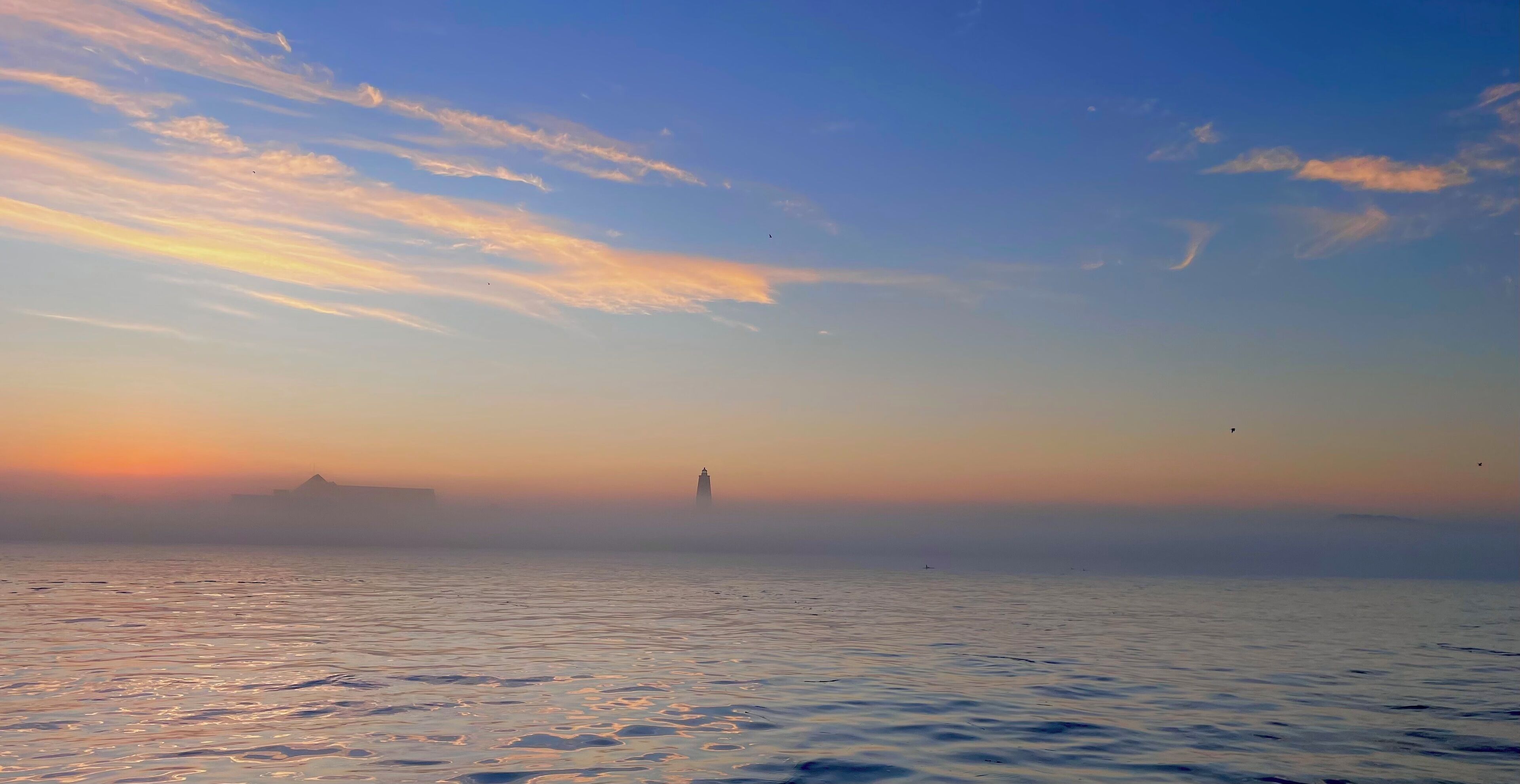 A calm sea at sunrise with a distant lighthouse and mist.
