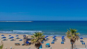 A beach with rows of blue lounge chairs and umbrellas, a lifeguard tower, and palm trees. - Ruby Studio - Old Town Rethymno (Rethymno)