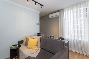 A modern living room with a sofa, a small side table, a coffee table, and a window with sheer curtains.