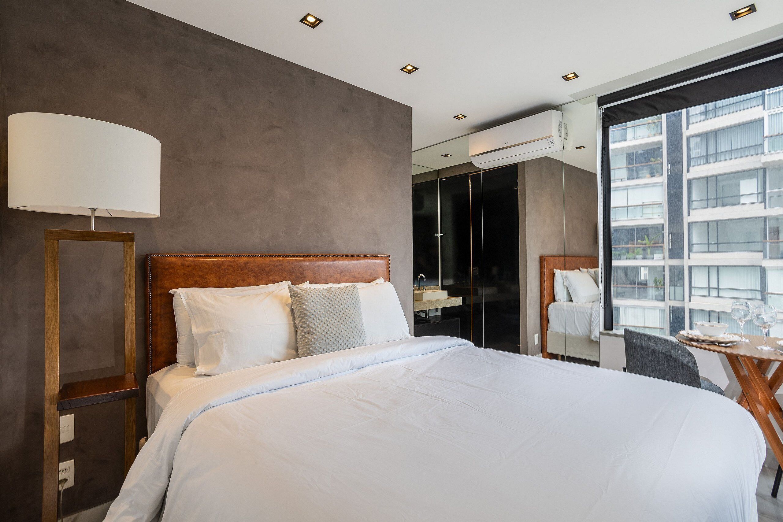 A modern hotel room with a large bed, a bedside table, a lamp, a small table with a chair, and a large window with a view of buildings.