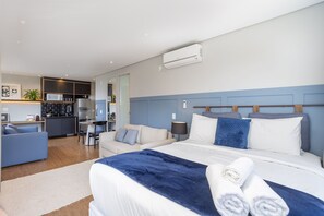 A modern hotel room with a bed, a sofa, a small kitchen area, and a dining table.