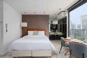 A modern hotel room with a large bed, a dining table with chairs, a city view, and a bathroom with a mirror and sink.