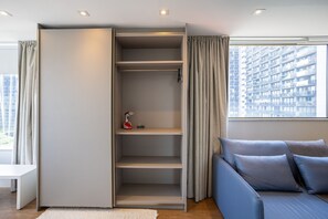 A modern room with a built-in closet, a sofa with cushions, a large window with curtains, and a view of a high-rise building.