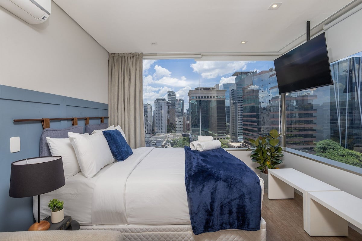 Een moderne hotelkamer met een groot bed, een televisie, uitzicht over de stad en een bankje bij het raam.
