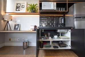 Une cuisine compacte équipée d’un four à micro-ondes, d’un réfrigérateur et de divers appareils électroménagers. On y trouve une étagère avec des objets décoratifs et une poubelle.
