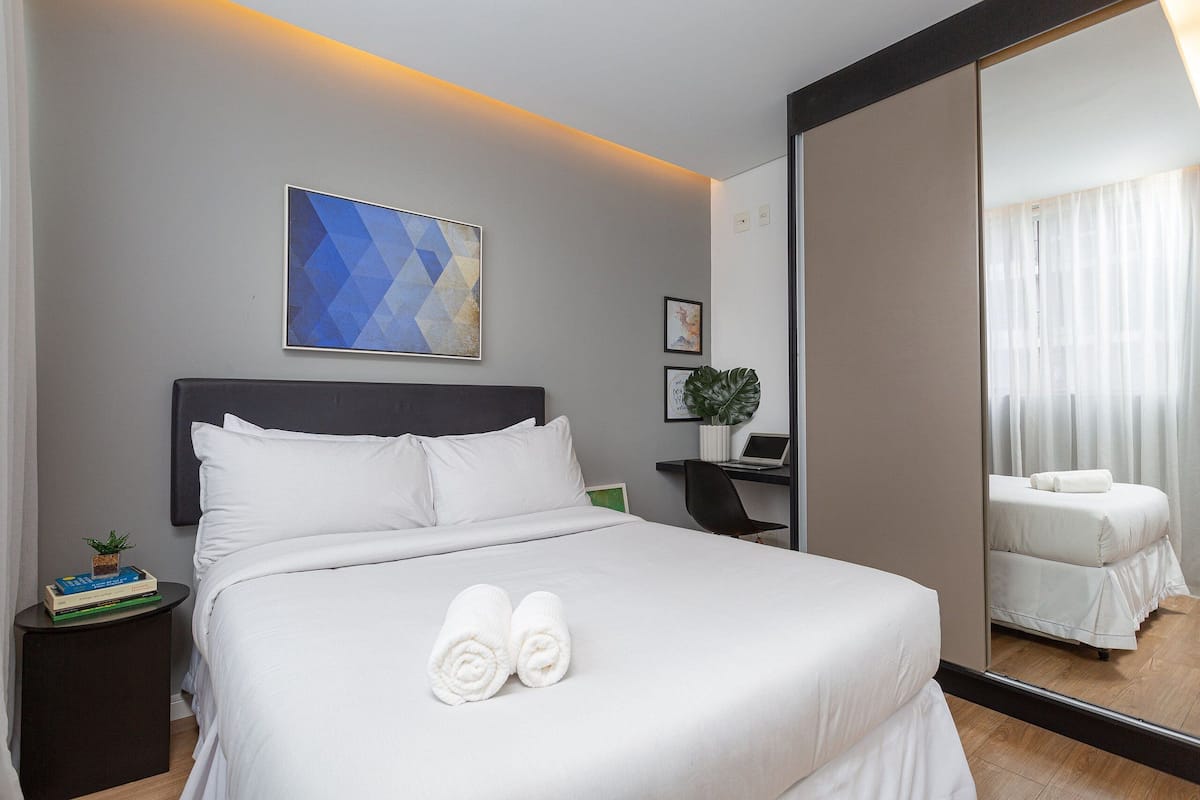 Een netjes opgemaakt bed met witte lakens en handdoeken, een klein nachtkastje met boeken en een plant, een bureau met een laptop, en een grote spiegel die de kamer weerspiegelt.