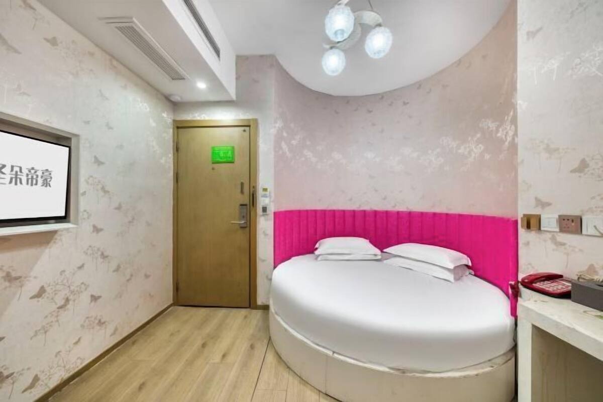 A hotel room with a round bed, a pink headboard, a wooden door, and a white wall with a screen displaying Chinese characters.