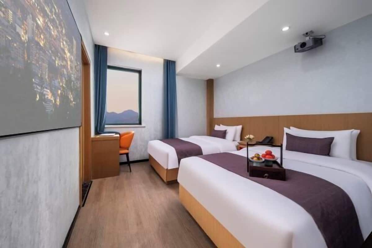 A hotel room with two beds, a desk, a chair, a projector screen, and a window with a view.