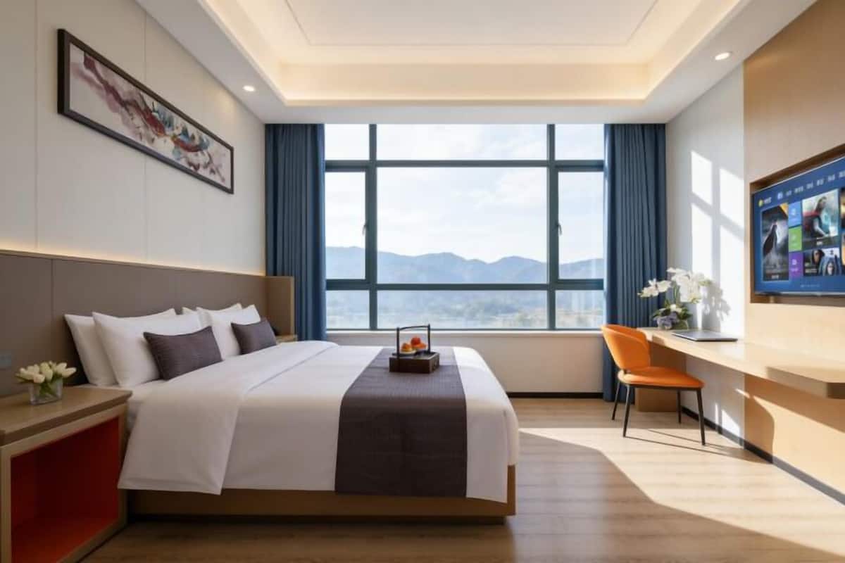 A hotel room with a bed, a desk, a chair, a window with a mountain view, and a painting on the wall.