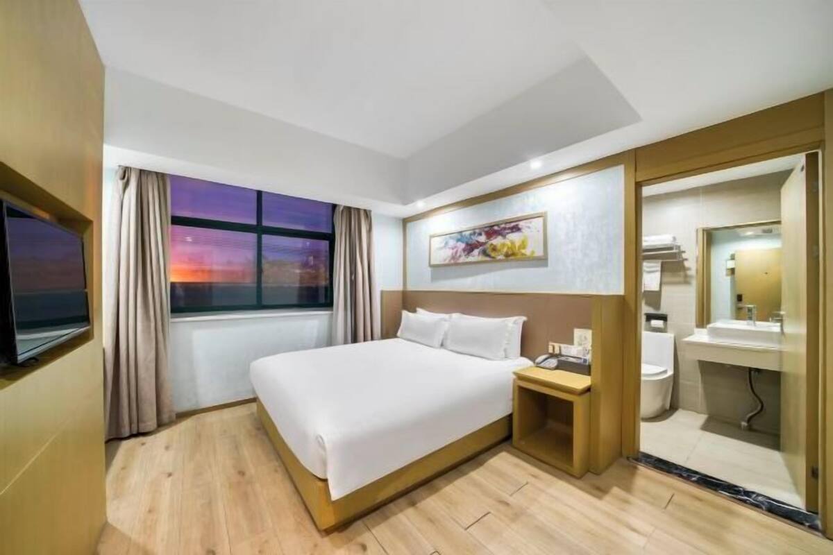A hotel room with a bed, a bedside table, a TV, a window with curtains, a painting, and a bathroom with a sink and mirror.