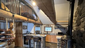 A kitchen with a central island, wooden bar stools, a stone wall, and a wood-burning stove.