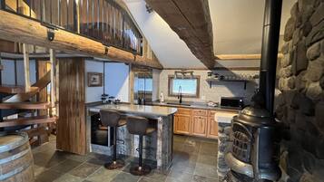 A kitchen with a central island, wooden bar stools, a stone wall, and a wood-burning stove.