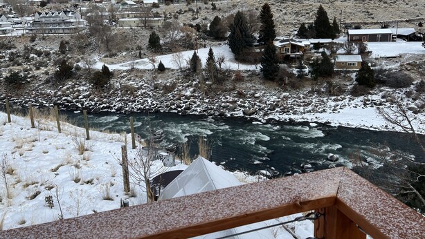 A snowy mountain landscape with a river in the foreground, surrounded by residential buildings and trees.