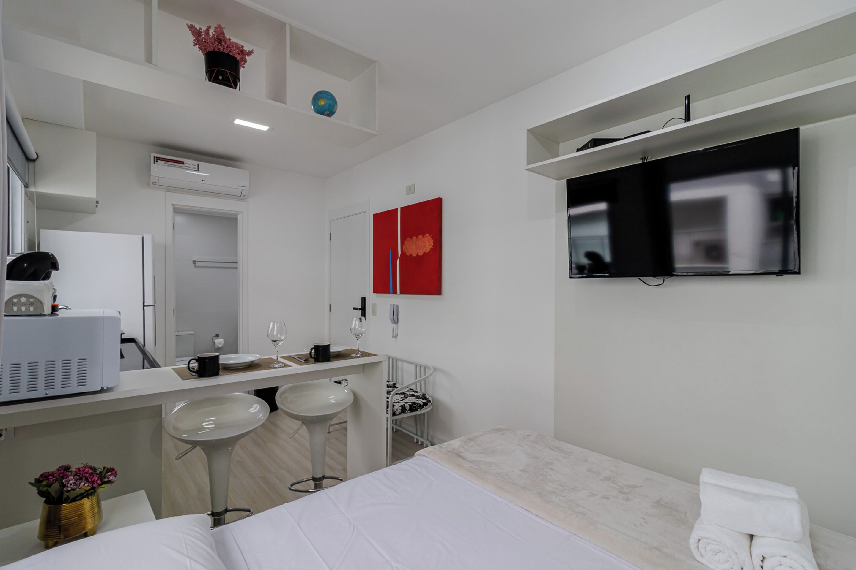 A modern hotel room with a bed, a TV mounted on the wall, a small bar area with stools, a sink, and a mirror.