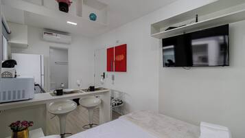A modern hotel room with a bed, a TV mounted on the wall, a small bar area with stools, a sink, and a mirror.