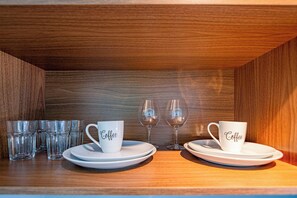 A wooden shelf with white coffee cups and plates, and two wine glasses.