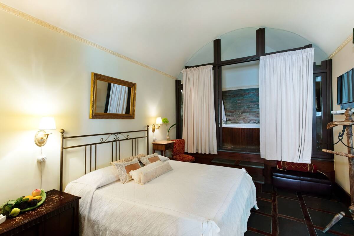 A hotel room with a bed, a nightstand with fruit, a lamp, a mirror, and a view of a brick wall through a window.