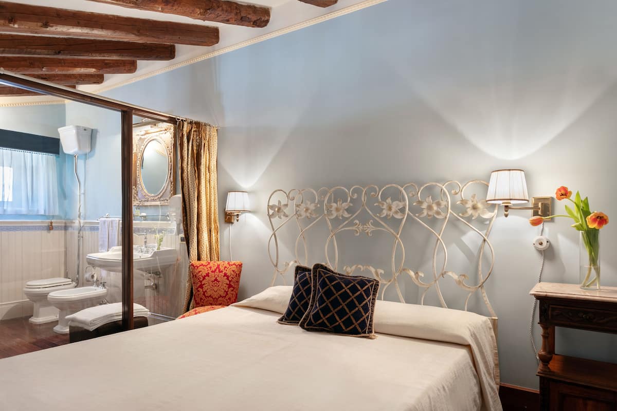 A bedroom with a bed, a nightstand, a mirror, and a bathroom visible through a glass door.